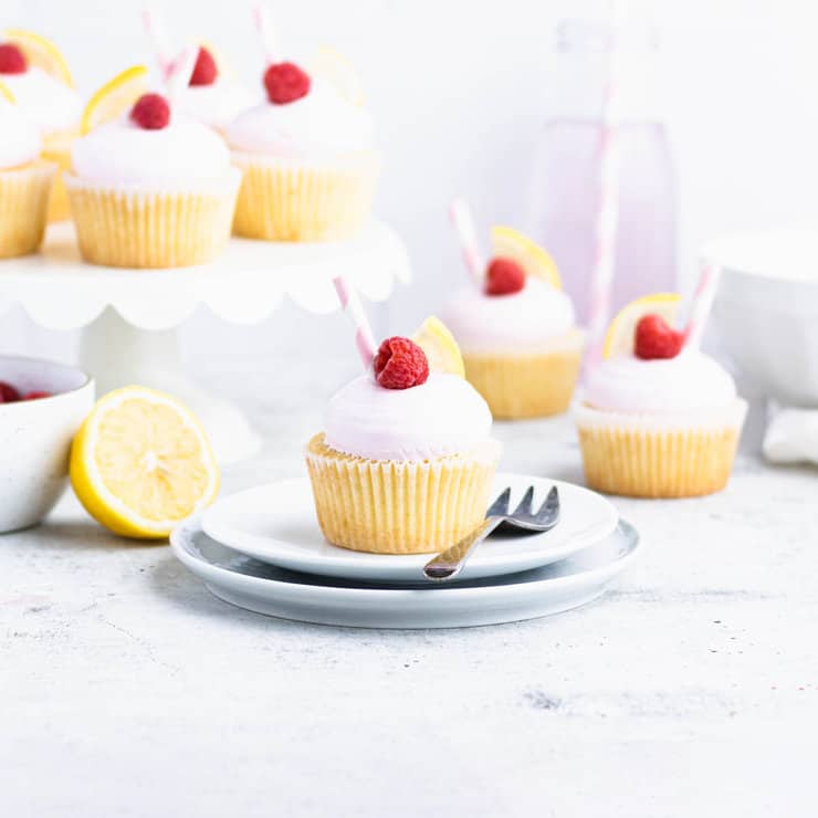 Yellow cupcake with pink frosting, a lemon wedge, raspberry and a pink striped straw on a plate. Other raspberry lemon cupcakes are in the background.