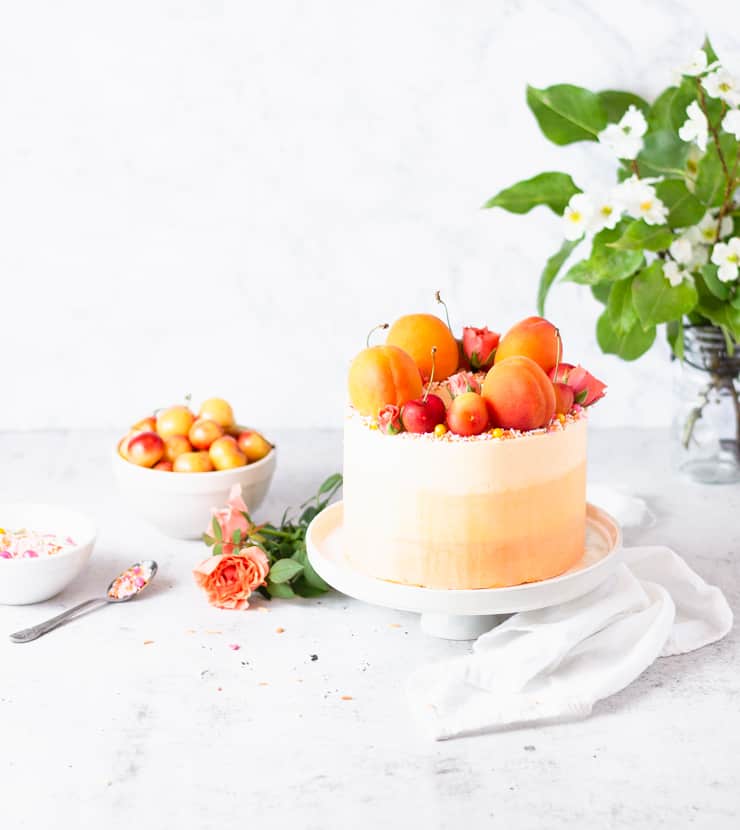 peach cake next on a table with cherries, sprinkles and a bouquet of flowers