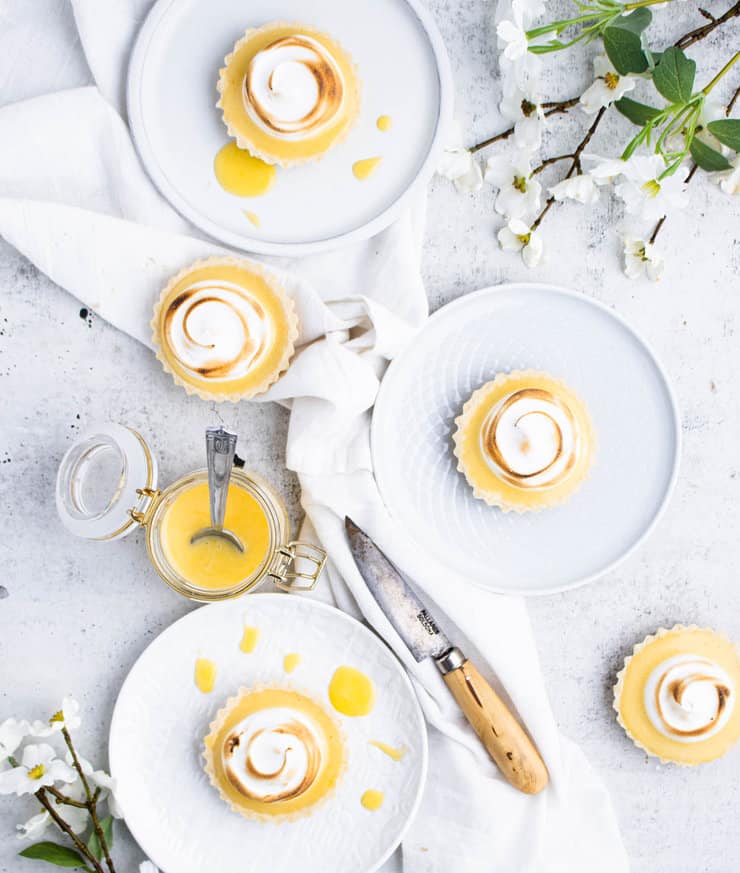 Small yellow tarts on  plates with a jar of lemon curd, a small paring knife and flowers around them.