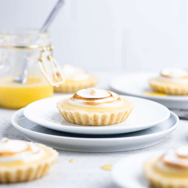 Lemon meringue tartlet on a white plate with other tarts and a jar of lemon curd around it.