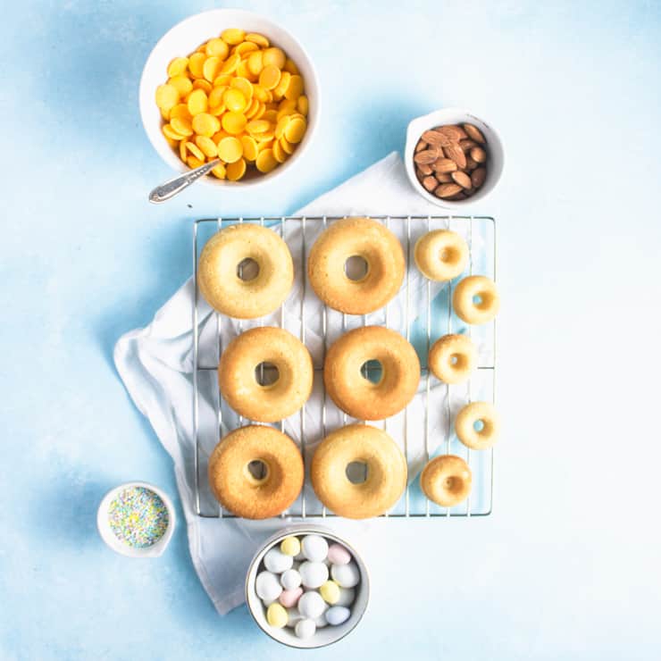Cooling rack with unfrosted donuts. Around the sides are a bowl of almonds, unmelted candy melts, sprinkles and egg candies.