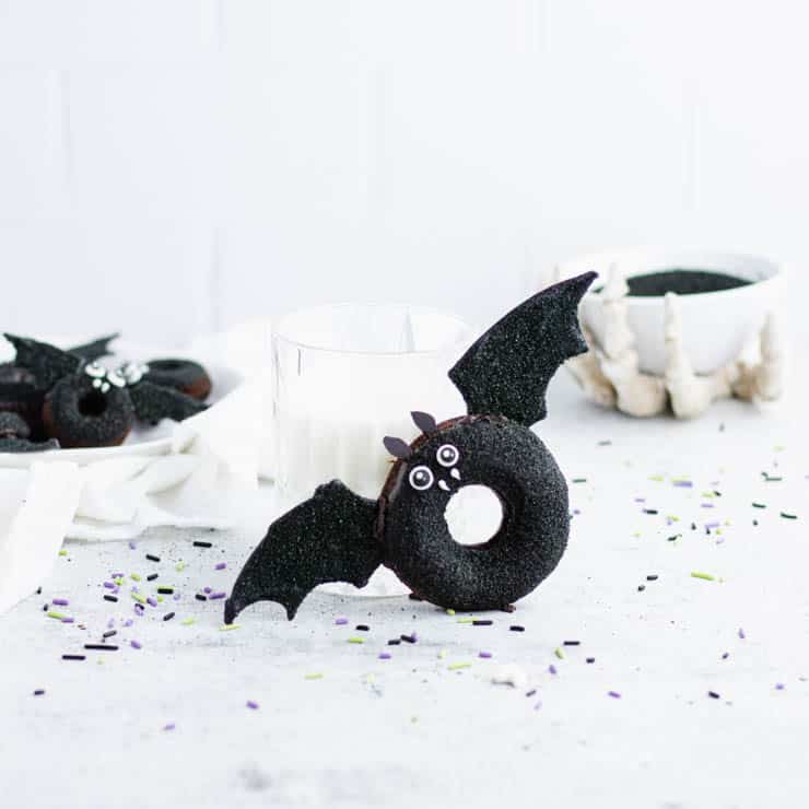 bat doughnut resting against a glass of milk with skeleton hand holding a white bowl and a plate of bat doughnuts in the background.