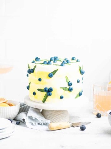 lemon blueberry poppy seed cake on a cake stand with drinks and plates around it.