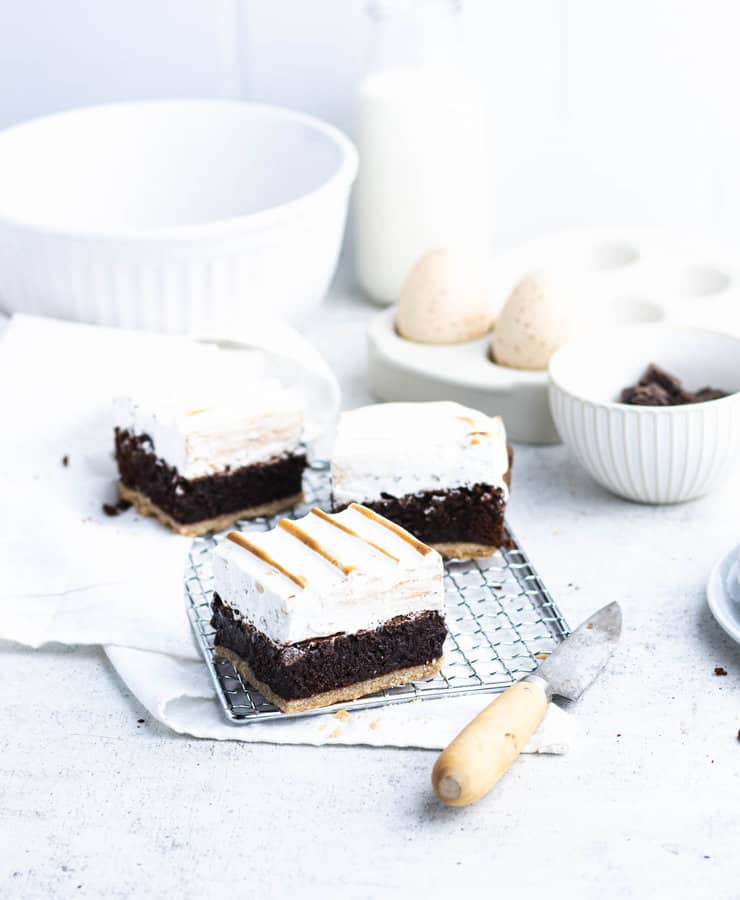 Three smore brownies on a silver cooling rack with two white bowls, a couple of eggs, a bottle of milk and a wooden handled knife in the background.