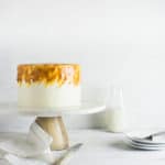 White frosted cake with caramel swirls on side against a white backdrop with a pitcher of milk and stacked plates
