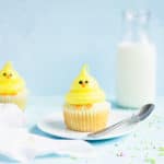 Chick cupcake on a plate with a spoon. A glass of milk is in the background with sprinkles strewn around the sides.