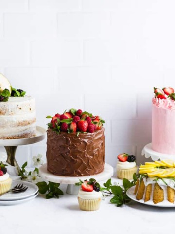 four cakes plus cupcakes lined up in front of a white tile backdrop