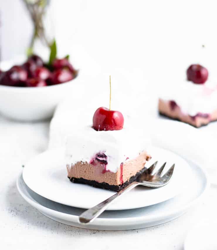 slice of black forest cheesecake on a white and blue plate placed at an angle so you can see layers of whipped cream, chocolate cheesecake and cherry compote.