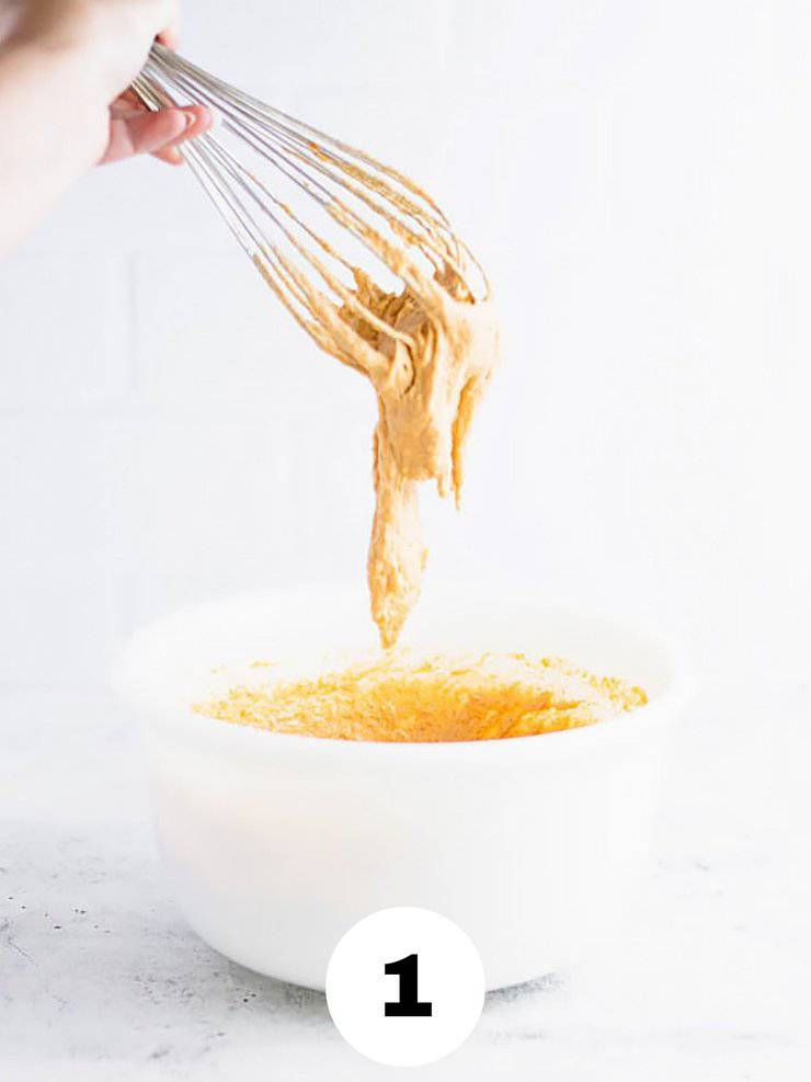 Whisk with thick, orange batter dripping off of it into a white bowl.
