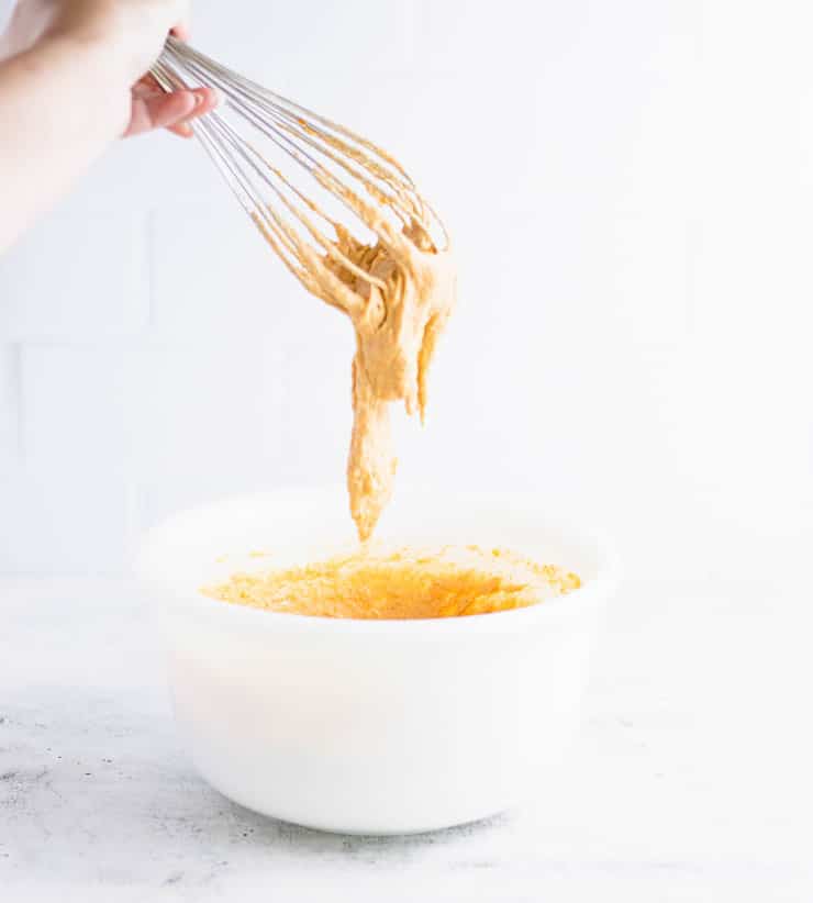 orange pumpkin waffle batter dripping into a white bowl from a copper colored whisk.