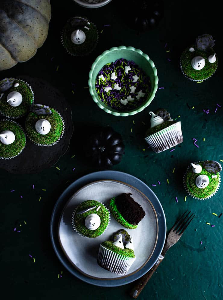 graveyard cupcakes on plates surrounding by sprinkles and off-white pumpkins against a dark green backdrop.