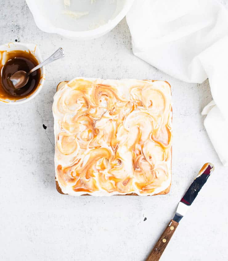 brown butter frosted apple cake with swirls of caramel next to a bowl of caramel sauce and an offset spatula with a wooden handle.