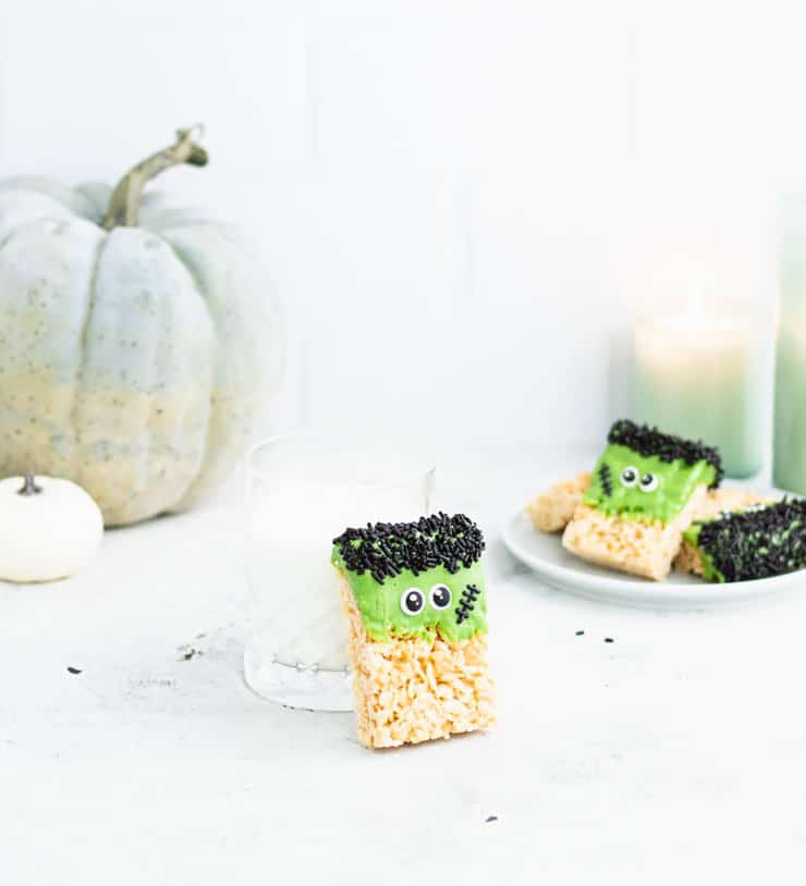 halloween rice krispie treat decorated with candy melts/sprinkles to look like frankenstein leaning against a glass of milk with a light green pumpkin, green candles and more rice krispie treats in the background.
