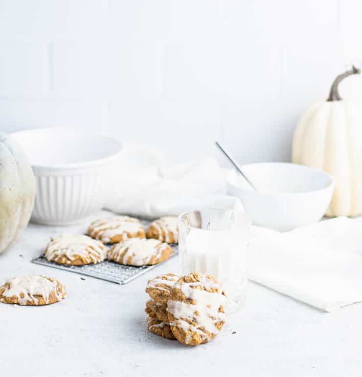 a stack of iced pumpkin cookies next to a short glass of milk with additional cookies, bowls and white and green pumpkins in the background against a tiled backdrop.