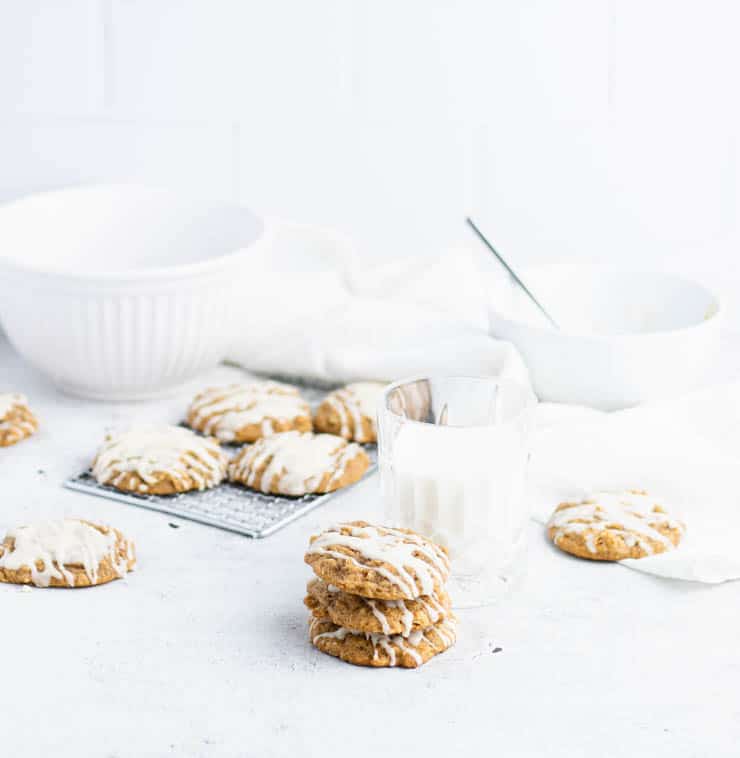 Iced pumpkin oatmeal cookies stacked in front of a cooling rack of cookies, and white bowls.