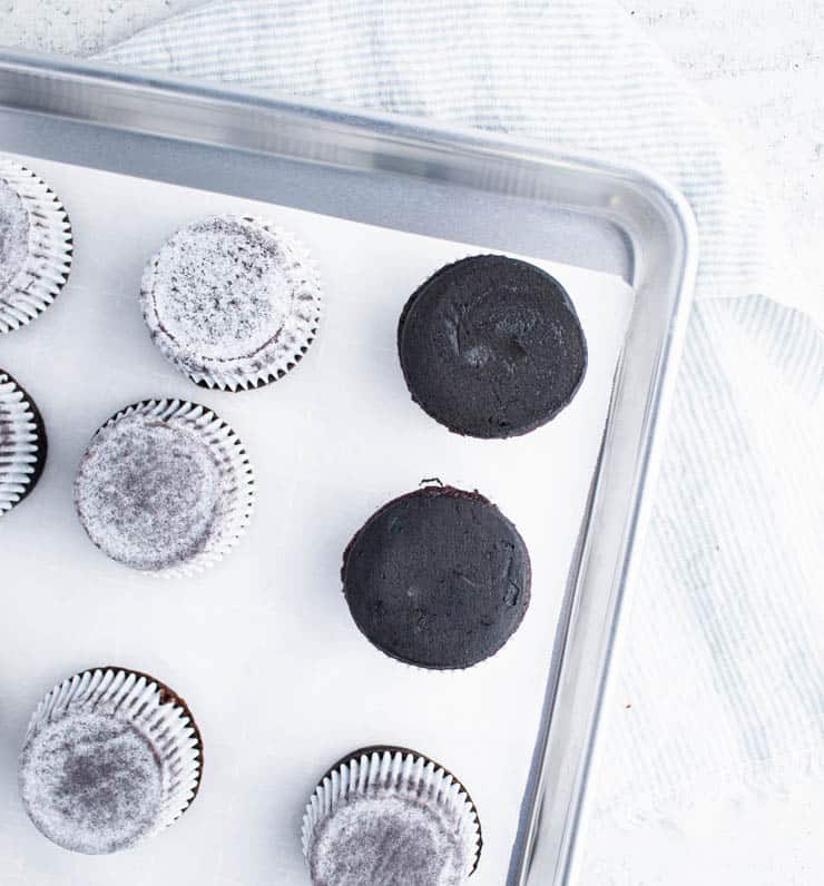 black frosted cupcakes frosting side down on a parchment paper lined silver baking sheet with two cupcakes turned upright to reveal a smoothly frosted top.