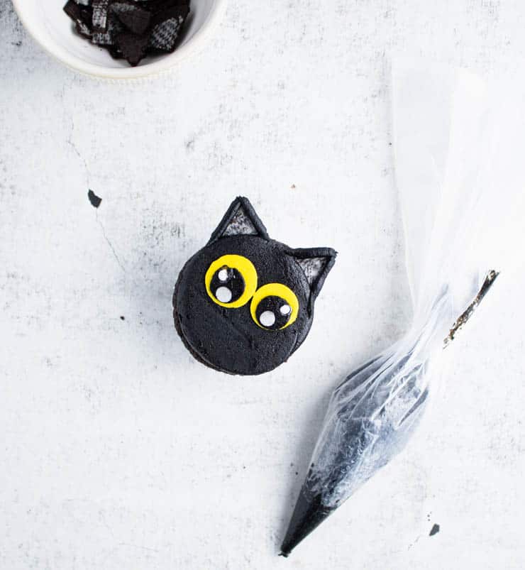 black cat cupcake with frosted ears next to a bag of black frosting and a bowl of Oreo cookies cut into triangles.