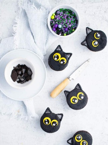 black cat cupcakes arranged against a white backdrop next to a bowl of sprinkles, a pairing knife with a light wood handle and a bowl of cut up Oreo cookies.