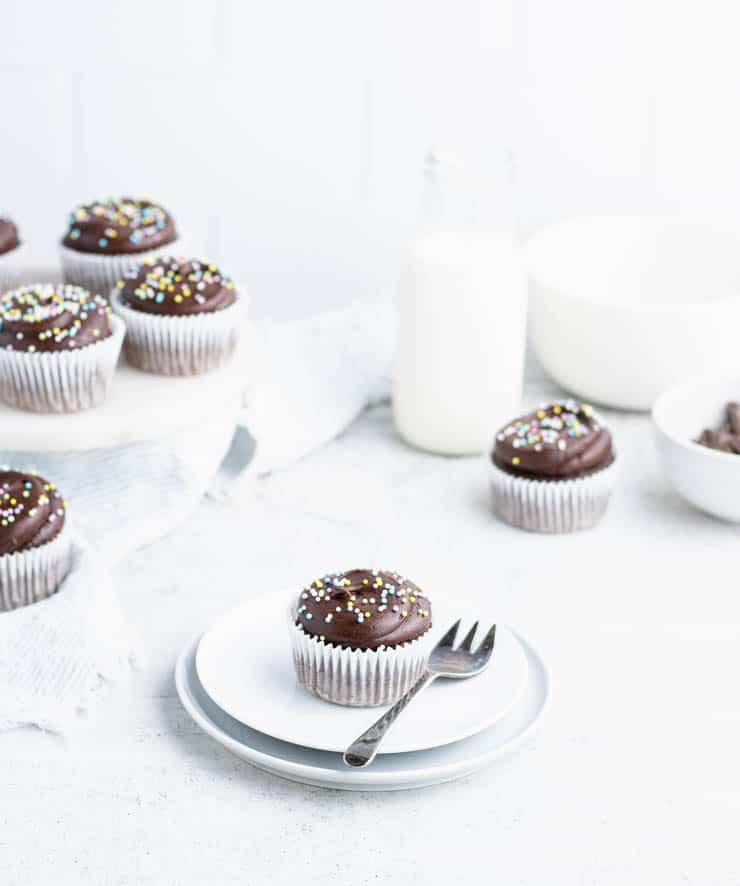 a chocolate frosted chocolate cupcake on a plate with other cupcakes and a glass of milk in the background.