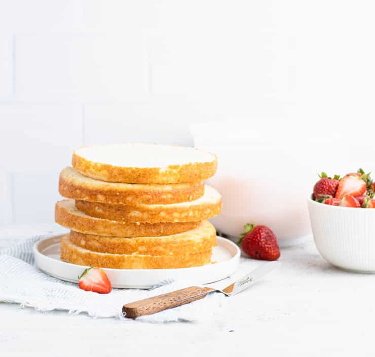 six layers of vanilla cake stacked on a plate with a bowl of strawberries and frosting in the background.