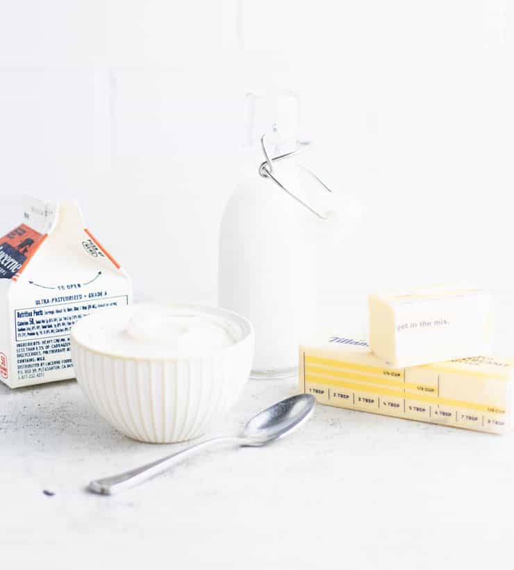 a small bottle of milk next to two sticks of butter, a bowl of yogurt and a small carton of cream.