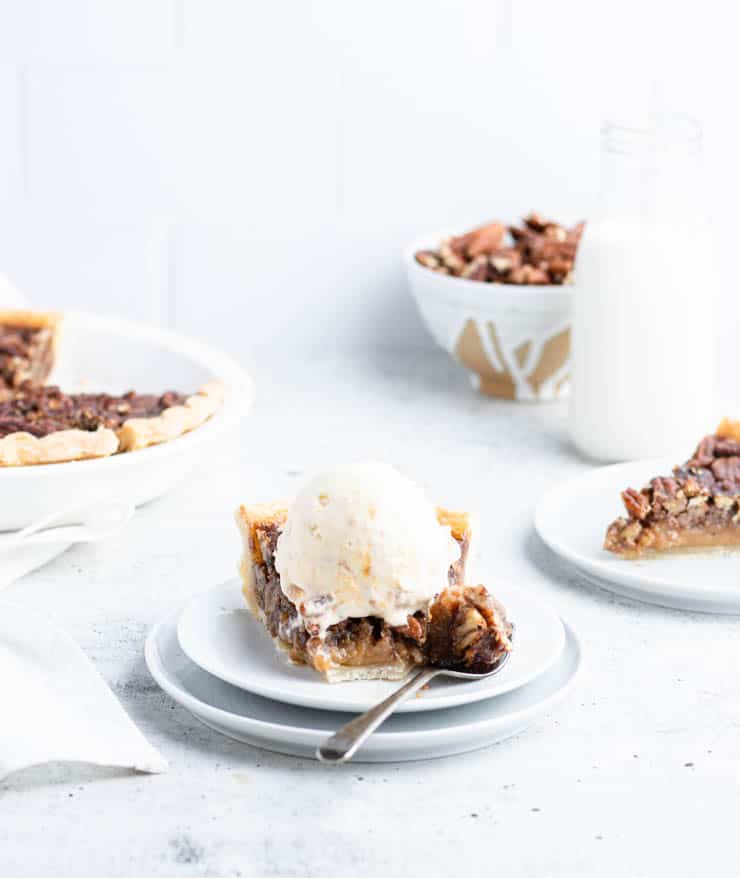 slice of bourbon pecan pie on a plate, topped with a scoop of ice cream and with a piece on a fork; In the background there's the whole pie, a bowl of pecans, a glass of milk and another plated slice.