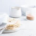 lefse stacked on a marble plate with a pitch, bowl and glass of cinnamon/sugar in the background.