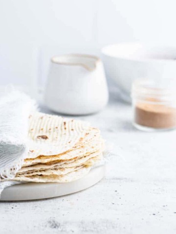 lefse stacked on a marble plate with a pitch, bowl and glass of cinnamon/sugar in the background.