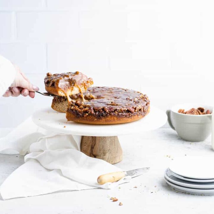 slice being pulled from an pecan upside down cake with bits of caramel pulling between the slice and the rest of the cake with plates and a bowl of pecans in the background.