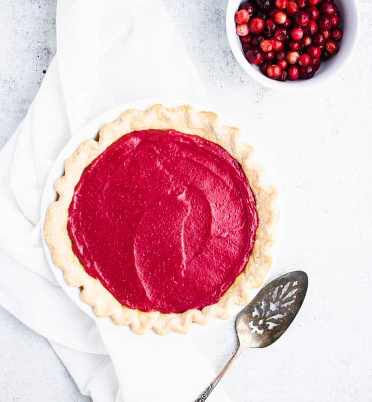 Bright red cranberry curd in a baked pie crust next to a spatula and a bowl of whole cranberries.