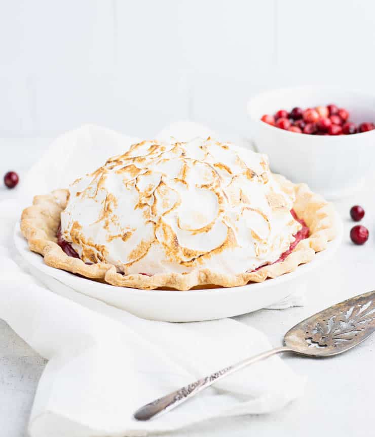 Bright red cranberry pie topped with a toasted mound of meringue in a white pie dish with a metal spatula in the foreground and a bowl of cranberries in the background.