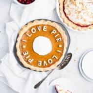 Pumpkin pie in a metal pie pan with cut out crust spelling out the phrase "I love pie" across the top. Empty plates, a bowl of cranberries and a cranberry pie are in the background.