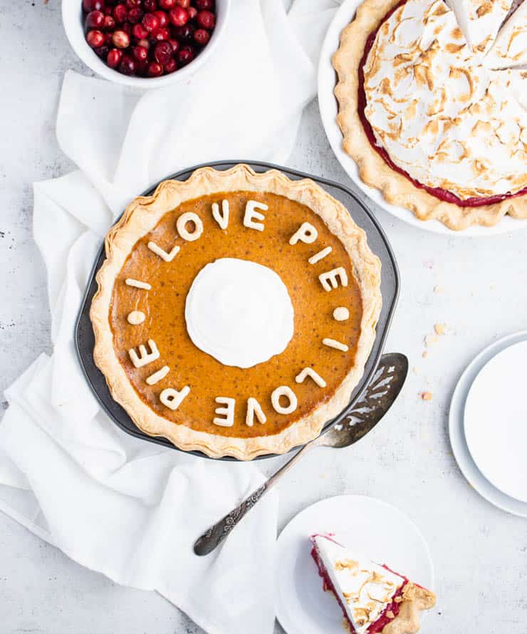 Pumpkin pie in a metal pie pan with cut out crust spelling out the phrase "I love pie" across the top. Empty plates, a bowl of cranberries and a cranberry pie are in the background.