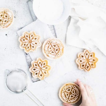 Overhead view of rosettes on a grey backdrop, some on a cooling wrack, some directly on the backdrop, with a hand reaching from the lower right side of the screen to take one.
