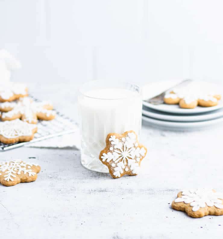 Iced snowflake shaped pepperkaker cookie resting against a glass of milk with other cookies in the background.