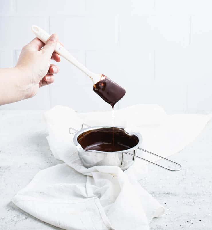Metal bowl of melted chocolate with a thin stream of chocolate dripping off of a spatula over the bowl.