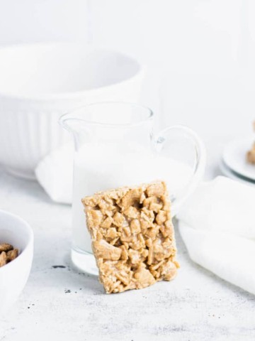 cinnamon toast crunch bar leaned against a small pitcher of milk with a bowl of cereal, a large white bowl and a plate of cereal bars in the background.