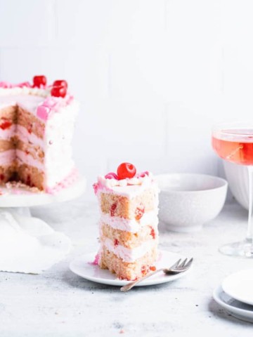 Slice of cherry chip cake with larger cake, white bowls and a cocktail in the background.