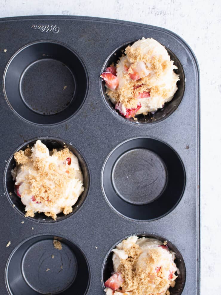 Strawberry muffin batter topped with brown sugar in a dark muffin tin.