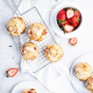 Overhead view of strawberry muffins on a small wire cooling rack with plated muffins, a bowl of strawberries and a bowl of brown sugar around it.