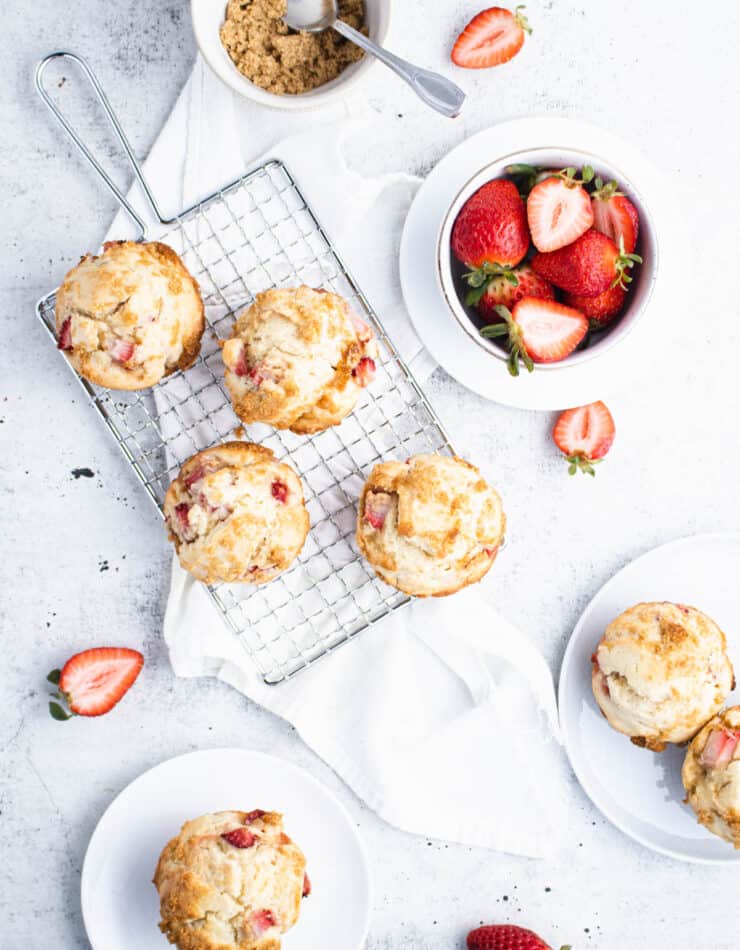 Overhead view of strawberry muffins on a small wire cooling rack with plated muffins, a bowl of strawberries and a bowl of brown sugar around it.