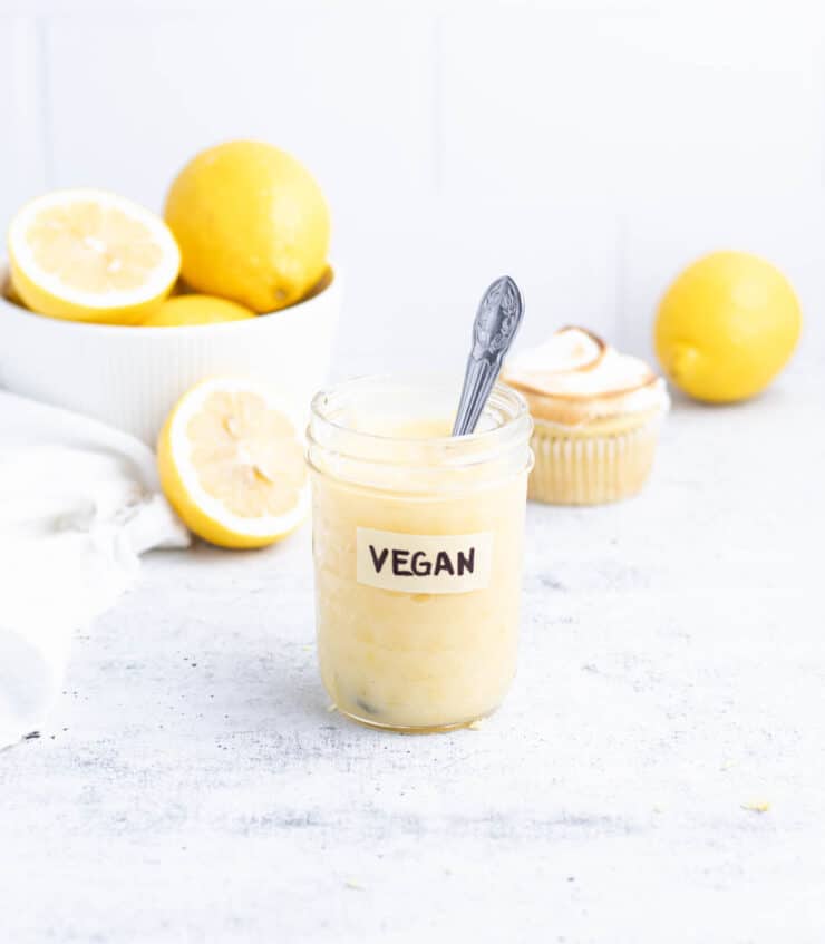 Jar of vegan lemon curd with a silver spoon in it. Bowl of lemons and a lemon meringue cupcake in the background.