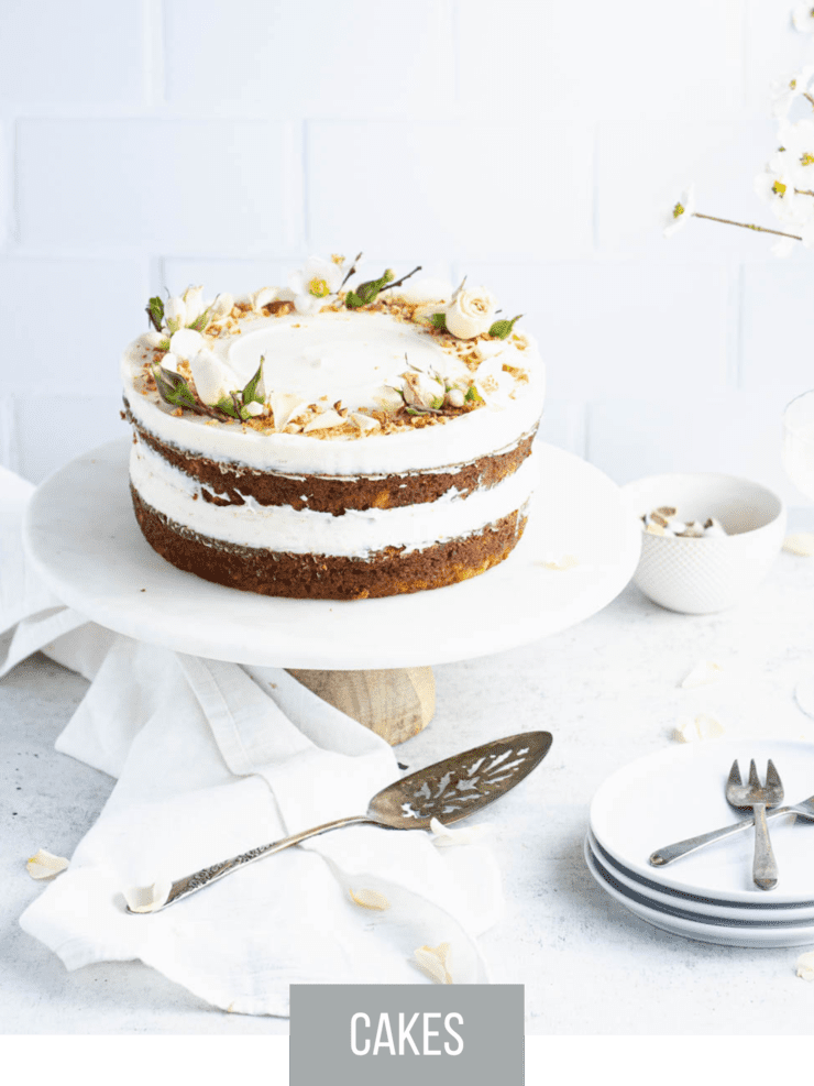 Pineapple carrot cake garnished with small pink flowers and chopped nuts on a marble cake stand with a light wood base. Plates, silverware and a bouquet of white flowers are in the background.