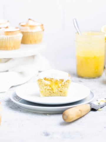 Halved lemon meringue cupcake with filling dripping out onto a white plate. Lemon curd, bowl of lemons and cupcakes in the background.