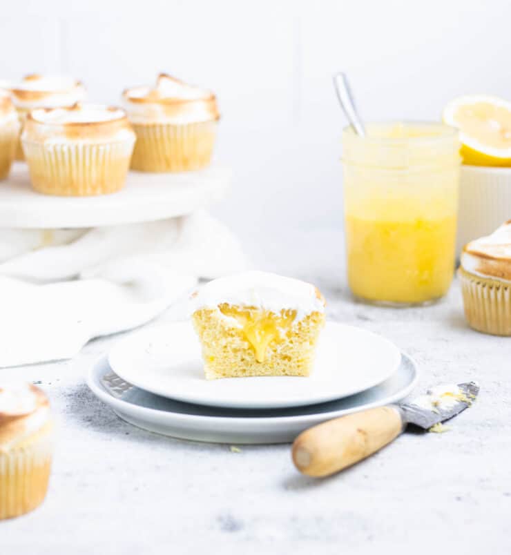 Halved lemon meringue cupcake with filling dripping out onto a white plate. Lemon curd, bowl of lemons and cupcakes in the background.