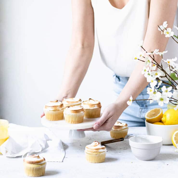 Person setting down a plate of lemon meringue cupcakes onto a white table. Cupcakes, bowl of lemons and white flowers in the foreground.