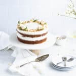 Pineapple carrot cake garnished with small pink flowers and chopped nuts on a marble cake stand with a light wood base. Plates, silverware and a bouquet of white flowers are in the background.
