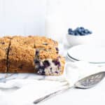 Side view of a square blueberry buckle with a sliced turned to show the inside of the cake. A bowl of blueberries, pitcher of milk and plates are in the background.