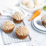 Cream cheese topped carrot cake muffins on a small silver cooling rack. Carrots, eggs, a white bowl and blue and white plates are in the background.