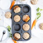 Overhead view of carrot cake muffins in a vintage slate colored muffin pan. Carrots, plates, silverware and other muffins are around it.